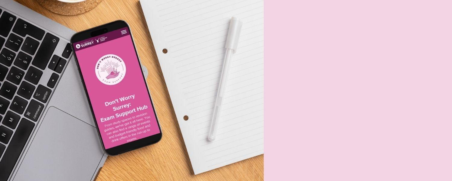 Flatlay of a phone showing the Don't Worry Surrey logo with a laptop and pen and paper