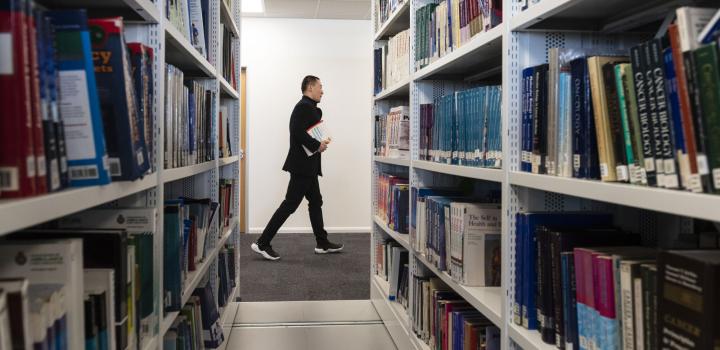 View through bookshelves filled with books, towards man walking from left to right at the end of the shelves, with a white wall in the background