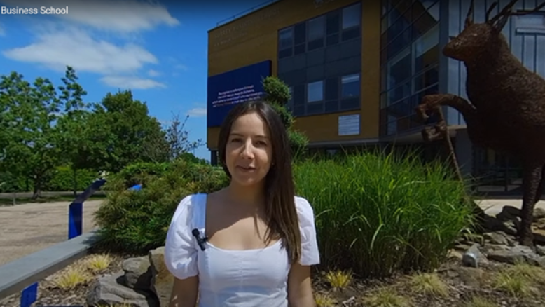 Still from video showing woman standing outside in front of plants and a building