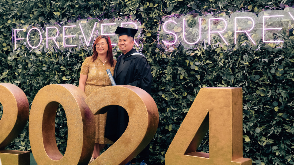 A graduate and their guest standing in front of a Forever Surrey sign and behind a 2024 sign at their graduation reception