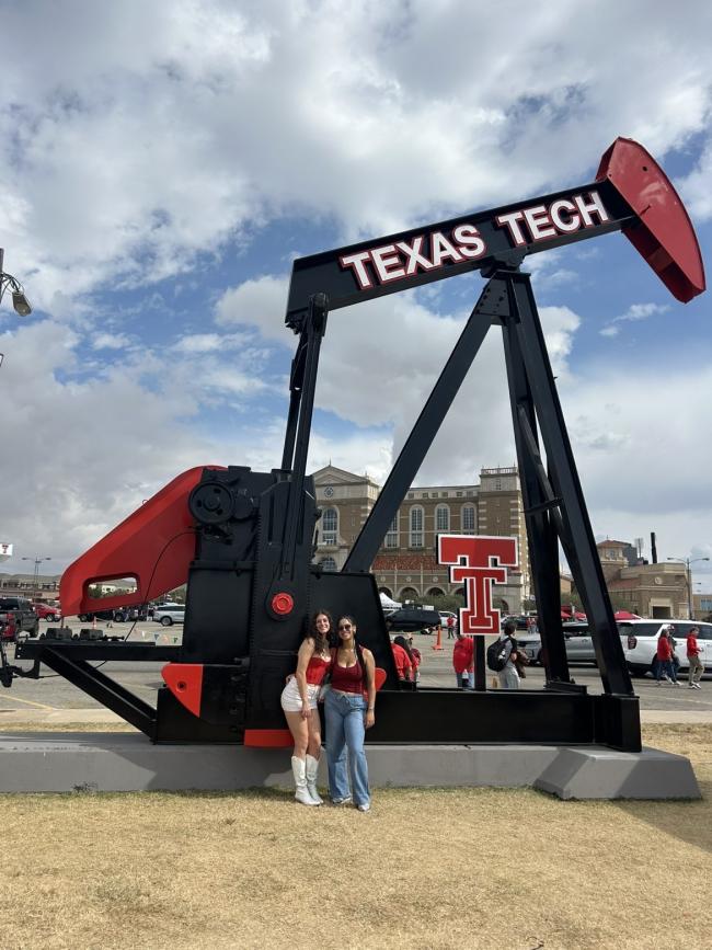 Two female students wearing red, standing in front of an oil pump with Texas Tech and the University logo.
