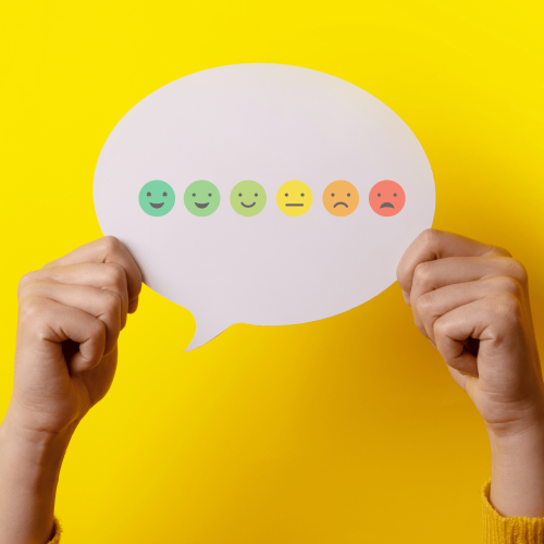 Two hands holding up a speech bubble containing emojis ranging from green happy to red unhappy. On a yellow background.